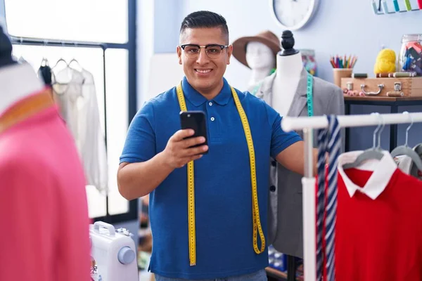 年轻的拉丁男子裁缝笑着自信地使用智能手机在工作室 — 图库照片