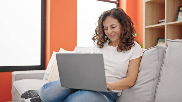 Латиноамериканка средних лет сидит дома на диване с ноутбуком