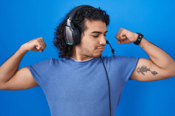 头发卷曲的西班牙裔男人用耳机听音乐时 露出胳膊肌肉的笑容 感到自豪 健身概念 — 图库照片