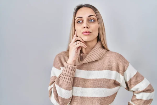 Young blonde woman wearing turtleneck sweater over isolated background with hand on chin thinking about question, pensive expression. smiling with thoughtful face. doubt concept.