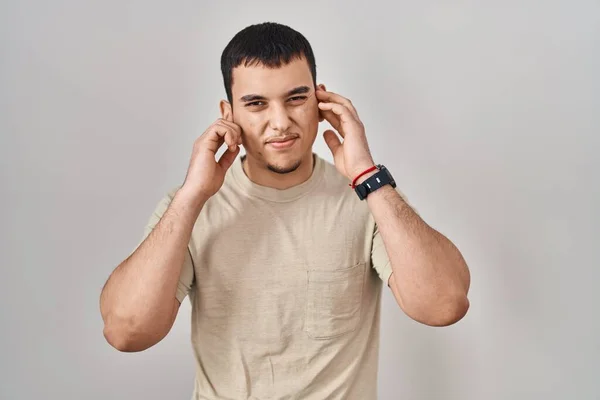 耳を指で覆っているカジュアルなTシャツを着た若いアラブ人男性は 大きな音楽のノイズに悩まされた表情をしています 聴覚障害の概念 — ストック写真