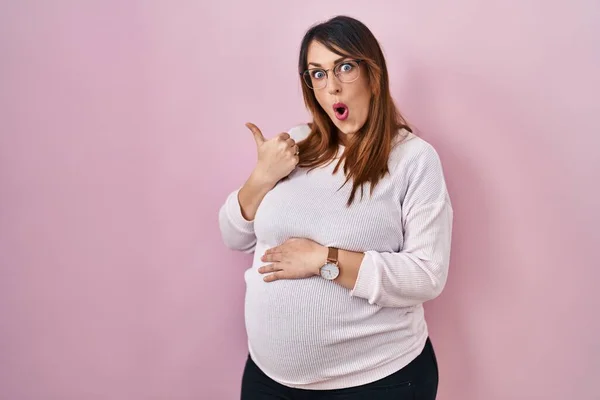 Těhotná Žena Stojící Nad Růžovým Pozadím Překvapeně Ukazuje Ukazováčkem Stranu — Stock fotografie