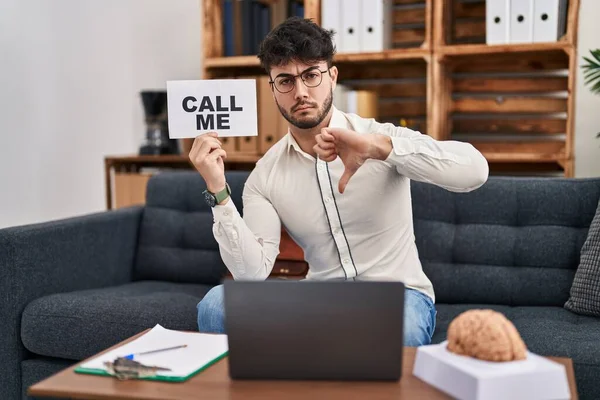 Hispanic man with beard working at therapy office holding call me sign with angry face, negative sign showing dislike with thumbs down, rejection concept