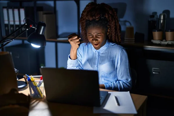 African woman working at the office at night angry and mad raising fist frustrated and furious while shouting with anger. rage and aggressive concept.