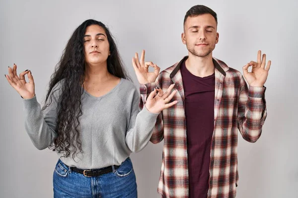 站在白色背景上的一对年轻的惊慌失措的夫妇放松了下来 闭上眼睛 用手指做冥想手势 瑜伽概念 — 图库照片