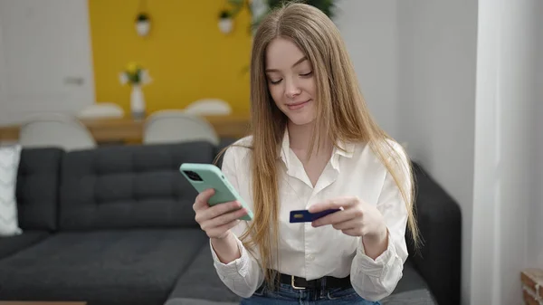 年轻的金发女人坐在家里的沙发上 手里拿着智能手机和信用卡购物 — 图库照片