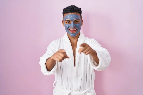 stock image Young hispanic man wearing beauty face mask and bath robe pointing to you and the camera with fingers, smiling positive and cheerful 