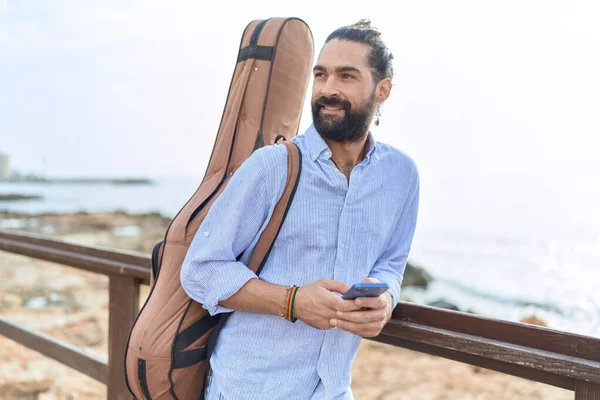 Young Hispanic Man Musician Using Smartphone Holding Guitar Case Seaside — Foto Stock