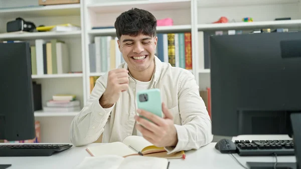 Young hispanic man student using smartphone sitting on table at library university