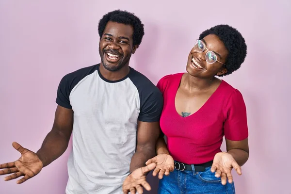 年轻的非洲裔美国夫妇站在粉色的背景上 高兴地微笑着 张开双臂表示友好的欢迎 积极的和自信的问候 — 图库照片