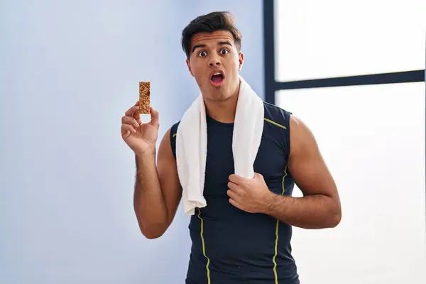 Hispanic man eating protein bar as healthy energy snack scared and amazed with open mouth for surprise, disbelief face