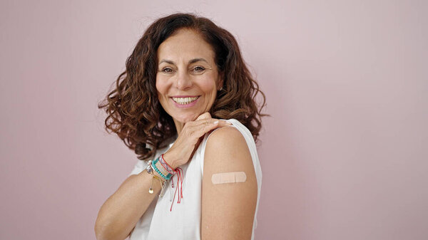 Middle age hispanic woman smiling confident standing with band aid on arm over isolated pink background