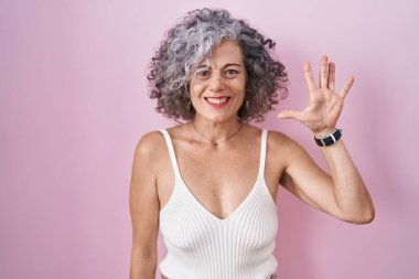 Middle age woman with grey hair standing over pink background showing and pointing up with fingers number five while smiling confident and happy. 