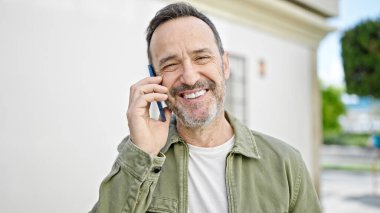 Orta yaşlı bir adam gülümsüyor. Sokakta akıllı telefondan konuşuyor.