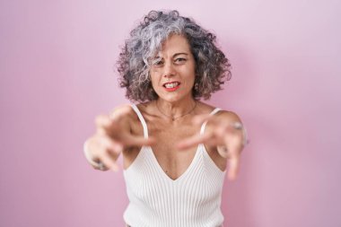 Pembe arka planda gri saçlı orta yaşlı bir kadın öfkeden kudurmuş, elleri boğmaya çalışıyor, deli gibi bağırıyor. 