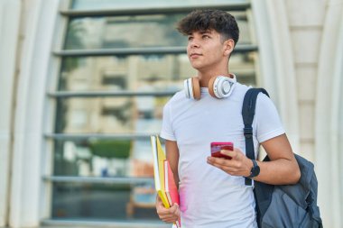 Young hispanic teenager student using smartphone holding books at university