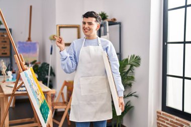 Sanat stüdyosundaki ikili olmayan kişi mutlu bir şekilde elini ve parmağını yana uzatarak gülümsüyor. 