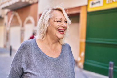 Orta yaşlı sarışın kadın sokakta kendinden emin bir şekilde gülümsüyor.