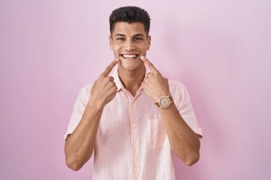 Young hispanic man standing over pink background smiling with open mouth, fingers pointing and forcing cheerful smile 