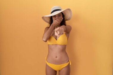 Bikini giyen ve yaz şapkası takan genç İspanyol kadın sana gülüyor, eli ağzında kamerayı işaret ediyor, utanç ifadesi var. 