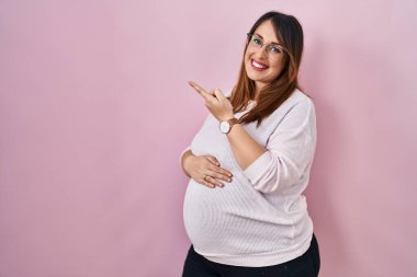 Pembe arka planda duran hamile bir kadın yüzünde kocaman bir gülümsemeyle elini kameraya doğru uzatarak işaret ediyor.. 