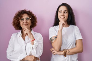 İspanyol anne ve kızı pembe arka plan üzerine günlük beyaz t-shirt giyiyorlar ve elleri çenelerinde soru sorup, düşünceli bir ifade takınıyorlar. Gülümseyen ve düşünceli bir yüz. şüphe kavramı. 