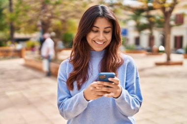 Genç İspanyol kadın parkta akıllı telefon kullanarak gülümsüyor.