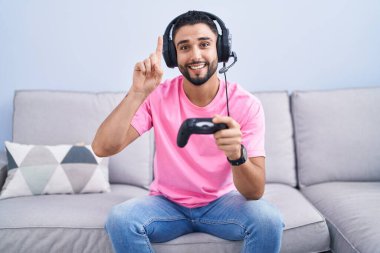 İspanyol genç adam video oyunu oynuyor kanepede oturmuş şaşkın şaşkın gülüyor, parmaklarıyla işaret ediyor ve kollarını kaldırıyordu.. 