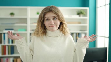 Young blonde woman student standing with confused expression at library university