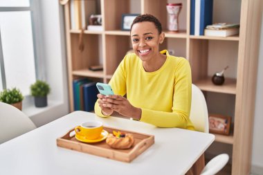 African american woman having breakfast using smartphone at home