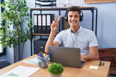 Ofiste çalışan genç İspanyol bir adam kulaklık takıyor. İki numaralı parmaklarıyla işaret ediyor ve gülümsüyor. Kendinden emin ve mutlu.. 