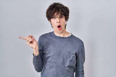 Young man standing over isolated background surprised pointing with finger to the side, open mouth amazed expression. 