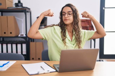 Ofiste çalışan genç İspanyol kadın gözlük takıyor ve gururla gülümseyen kol kaslarını gösteriyor. fitness konsepti. 