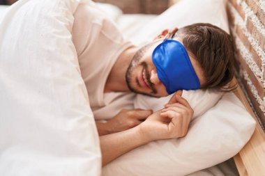 Uyku maskesi takmış genç beyaz adam yatak odasında uyuyor.
