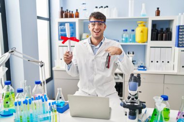 Laboratuvarda çalışan Latin asıllı genç bir adam diplomasını elinde tutarak mutlu ve gururlu gülümseyen birini işaret ediyor. 