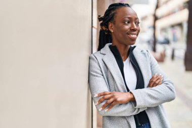 Afro-Amerikalı kadın gülümsüyor. Kollarını kavuşturup caddede duruyor.