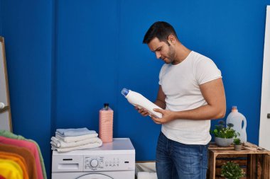 Young hispanic man holding detergent bottle at laundry room