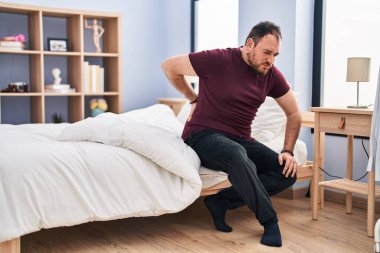 Young hispanic man suffering for backache sitting on bed at bedroom