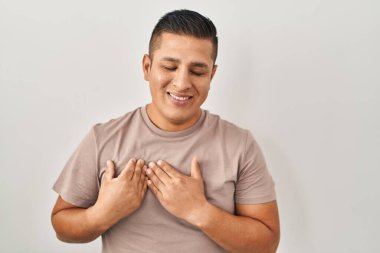 Beyaz arka planda duran İspanyol genç adam elleri göğsünde, gözleri kapalı ve yüzünde minnet dolu bir jest ile gülümsüyor. sağlık kavramı. 