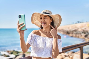 Genç Brezilyalı bir kadın akıllı telefonuyla fotoğraf çekiyor açık havada gülümsüyor mutlu bir şekilde eli ve parmağı ile işaret ediyor 