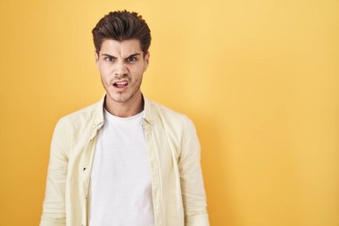 Young hispanic man standing over yellow background in shock face, looking skeptical and sarcastic, surprised with open mouth 