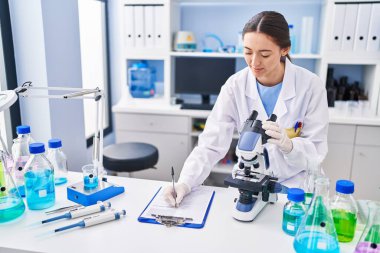 Young beautiful hispanic woman scientist using microscope writing on document at laboratory