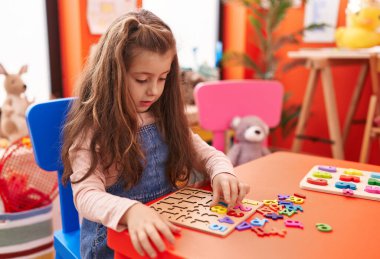Adorable hispanic girl playing with maths puzzle game sitting on table at kindergarten