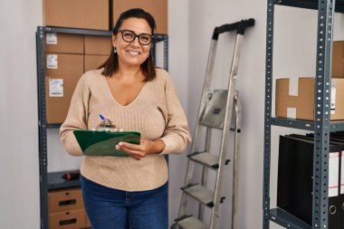 Middle age hispanic woman ecommerce business worker writing on clipboard at office