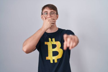 Beyaz, sarışın, bitcoin tişörtlü bir adam sana gülüyor, kamerayı işaret ediyor, ağzı kapalı, utanç ifadesi var. 