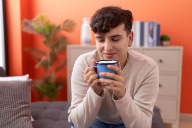 Non binary man drinking coffee sitting on sofa at home