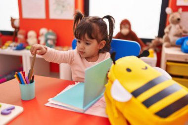 Anaokulunda elinde renkli kalemlerle masada oturan sevimli İspanyol kız öğrenci.