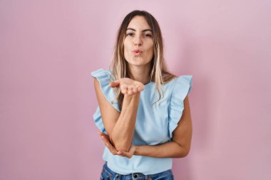 Young hispanic woman standing over pink background looking at the camera blowing a kiss with hand on air being lovely and sexy. love expression. 