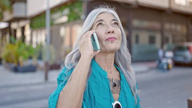 Orta yaşlı, kır saçlı, akıllı telefonlu bir kadın sokakta ciddi bir ifadeyle konuşuyor.