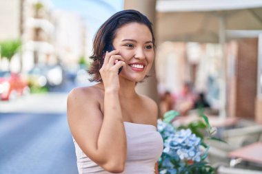 Genç İspanyol kadın gülümsüyor. Kafe terasında akıllı telefondan konuşuyor.